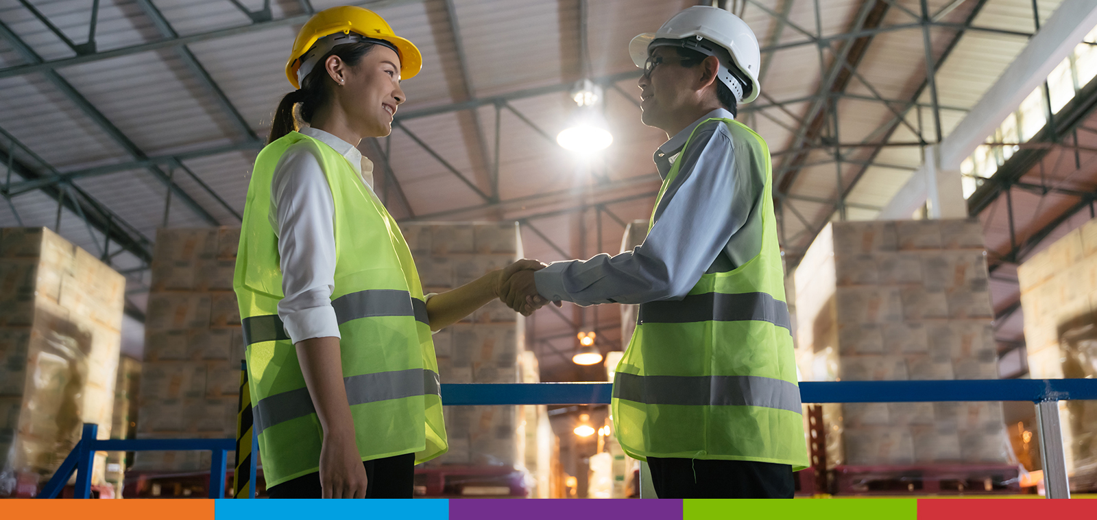 Une femme portant un casque jaune serrant la main d'un homme portant un casque blanc dans une usine pleine de palettes de boîtes